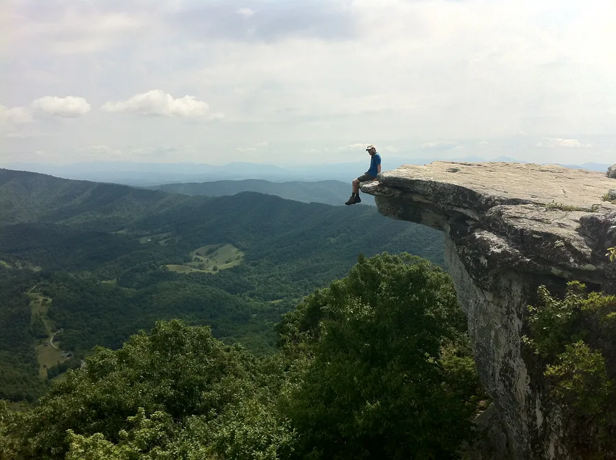 A hiker sitting on the edge of a rock ledge above forested Virginia valleys